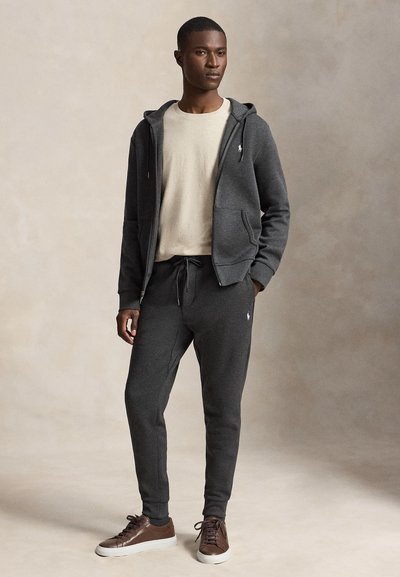 Άνδρας που φοράει σκούρο γκρι hoodie με φερμουάρ και αντίστοιχο φόρμας, μπεζ μπλουζάκι και καφέ αθλητικά παπούτσια, στέκεται μπροστά σε ουδέτερο φόντο.