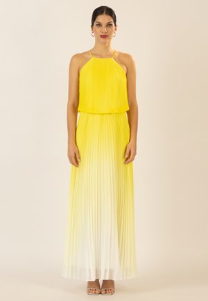 Frau steht und trägt ein ärmelloses langes Maxikleid mit gelb-weißem Ombre-Effekt und dünnen Trägern vor einem schlichten beigen Hintergrund.