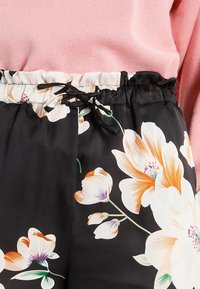 Pantalon noir à motifs floraux blancs et orange, doté d'une ceinture élastique avec cordon de serrage et d'une texture satinée lisse.