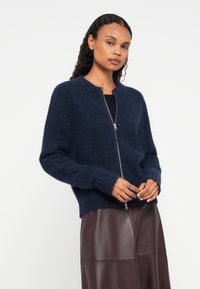 Moteris su garbanotais plaukais vilkinti tamsiai mėlyną pūkuotą džemperį su užtrauktuku ir tamsiai rudą odinę sijoną prieš paprastą baltą foną.