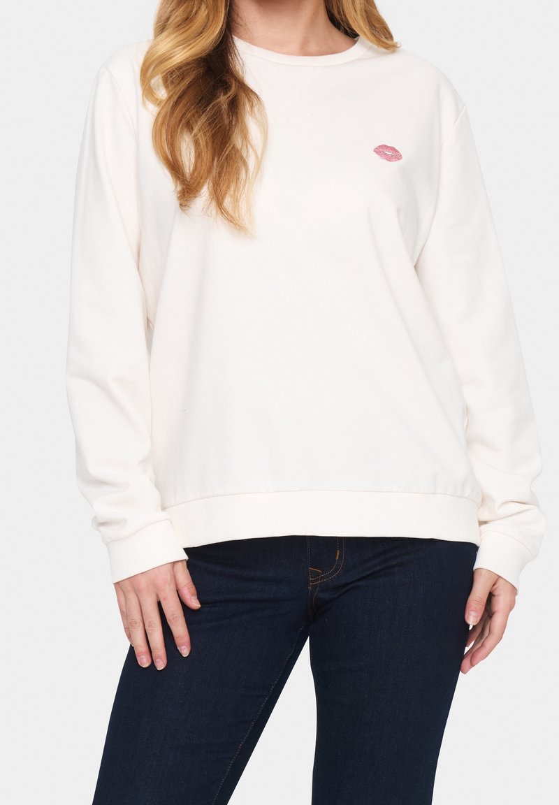 Femme portant un sweat-shirt blanc avec une petite empreinte de lèvre rose sur la poitrine et un jean bleu foncé, devant un fond uni.