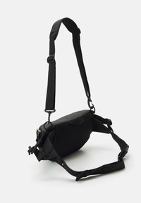 Svart justerbar crossbody-väska med mesh-bakdel, zippered fack, spännen och vadderad axelrem på vit bakgrund.