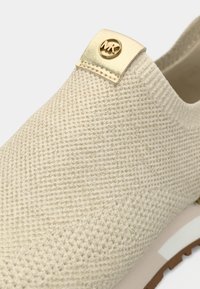 Beige liukusukenkä, jossa on teksturoitu, kudottu materiaali, kultainen logokoriste kantapäässä ja paksu, vaaleanvärinen pohja.