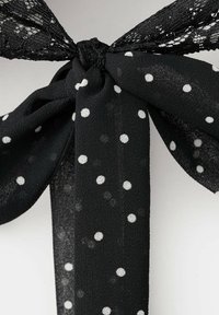 Nœud en tissu noir avec un côté en dentelle et l'autre en tissu à pois, noué en un nœud lâche sur un fond gris.