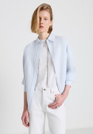 Woman with short blonde hair wearing a blue striped button-up shirt over a white top, white pants, silver ring, and necklace, standing against white background.