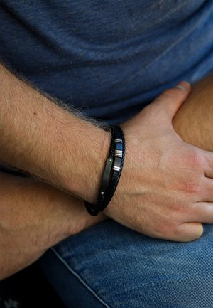 Schwarzes Lederarmband mit geflochtenem Muster und metallischen Akzenten, ausgestattet mit einem polierten Verschluss. Am Handgelenk getragen, vor einem blauen Textilhintergrund.