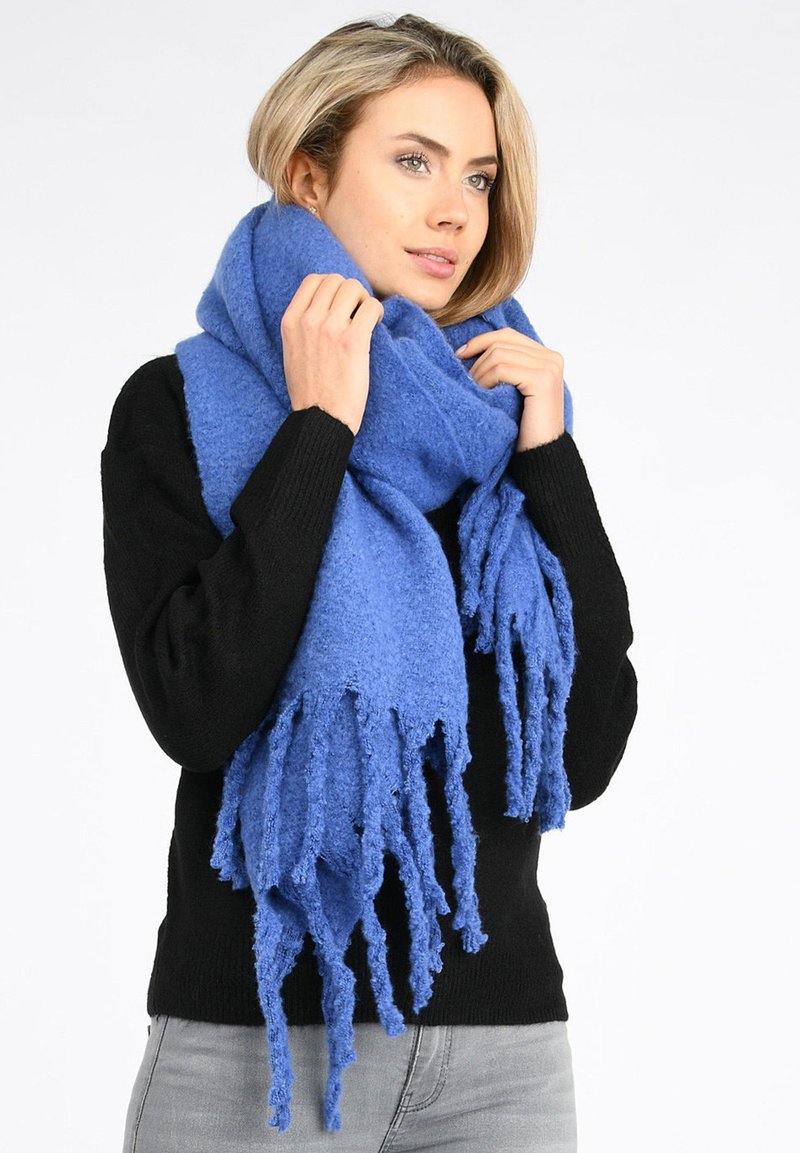 Jeune femme portant une grande écharpe bleue à franges et un pull noir, ajustant l'écharpe autour de son cou, fond blanc.