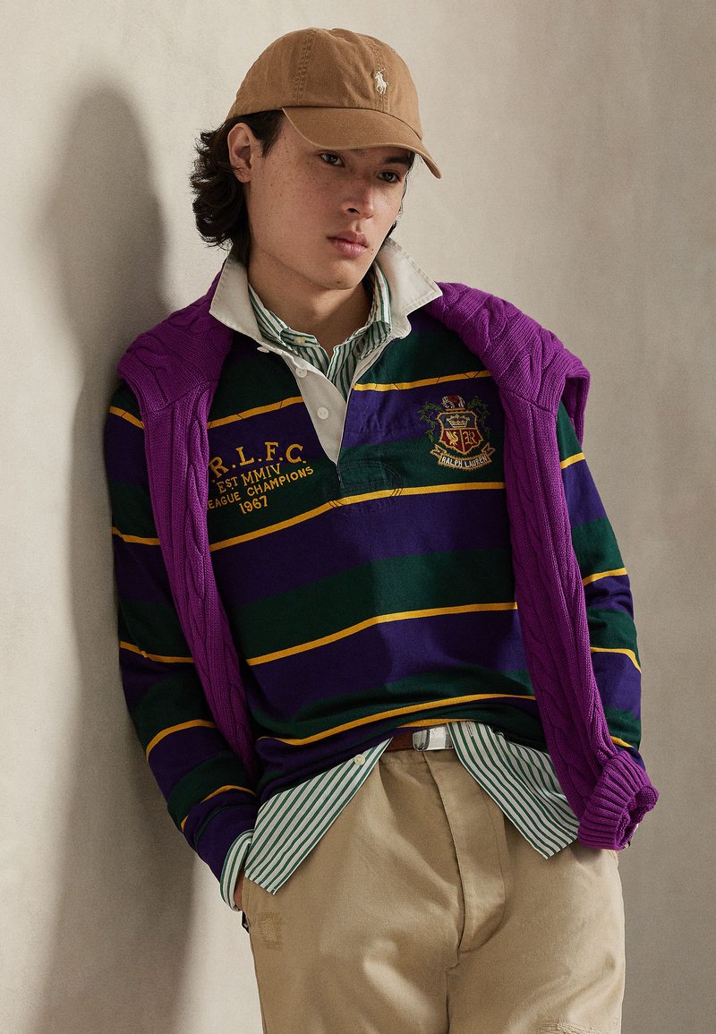 Pull violet à câbles drapé sur un maillot de rugby rayé vert et violet avec des accents jaunes, porté avec un pantalon beige et une casquette marron clair.