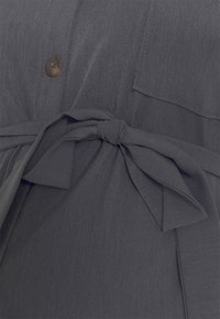 Šedá blůza s texturovanou tkaninou, předním zapínáním na knoflíky, dvěma kapsami na hrudi a detailem vázání v pase pro úpravu.