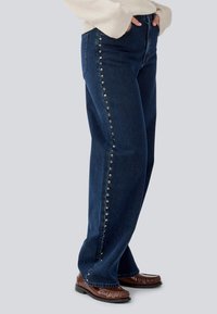 Dark blue denim jeans with a straight leg design, featuring silver snap buttons along the side, and a high waistband.