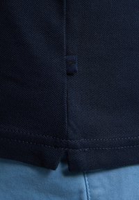 Mörkblå polotröja med texturerad tyg, med en sidoklyvning och en liten logotagg i fållen. Kombineras med ljusblå byxor.