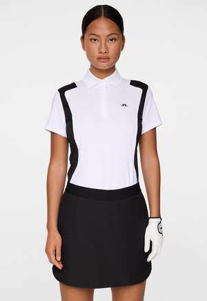 Donna che indossa una polo da golf bianca e nera, una gonna nera e un guanto da golf bianco sulla mano sinistra su uno sfondo neutro.