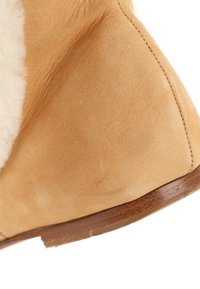 Gros plan d'une chaussure en daim beige avec doublure en shearling crème et semelle en bois marron, mettant en valeur les coutures et les détails de la texture.