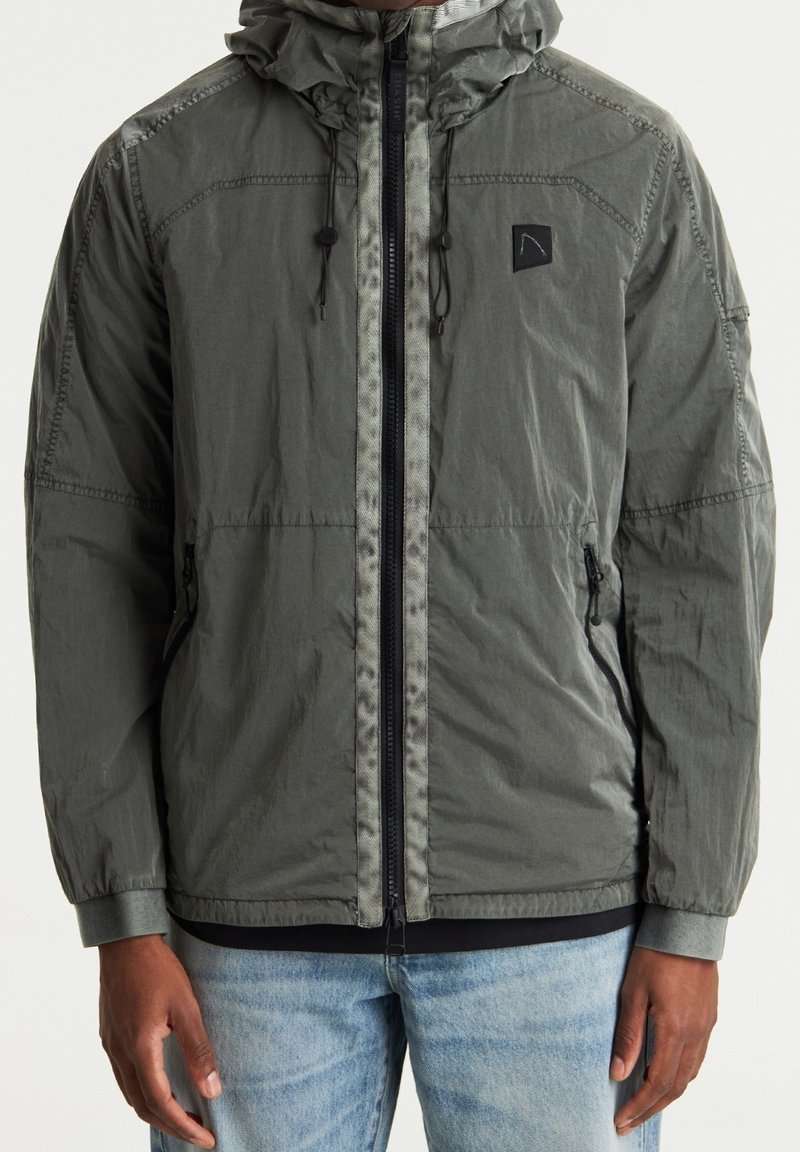 Lekki płaszcz z kapturem w oliwkowym kolorze, z pełnym zamkiem błyskawicznym, elastycznymi mankietami i dwiema bocznymi kieszeniami. Tkanina ma subtelny połysk i fakturę.