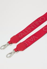Correia de tecido vermelho com logótipo em relevo "Valentino", apresentando dois clips metálicos e acabamentos em pele em ambas as extremidades. Textura suave e design robusto.