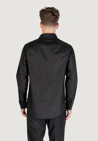 Camicia nera a maniche lunghe con un motivo a diamante testurizzato sulla schiena, polsini con bottoni e colletto classico. Finitura in tessuto liscia.