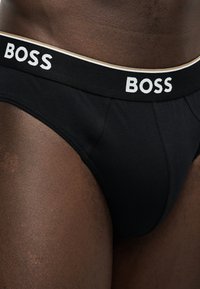 Close-up of a person wearing black briefs with a waistband labeled BOSS in white letters on a dark skin tone.