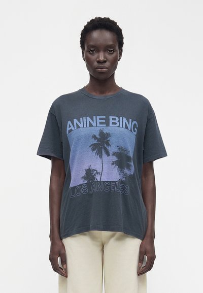 Άτομο που φοράει σκούρο φαρδύ T-shirt με μπλε φοίνικες και το κείμενο "ANINE BING" και "LOS ANGELES", σε συνδυασμό με ανοιχτόχρωμο παντελόνι.