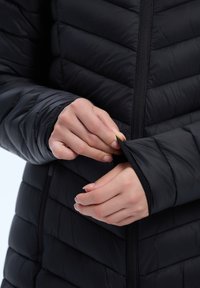 Mustavillinen eristetty takki, jossa on tyylikäs tikkikuvio. Lähikuva käsistä, jotka vetävät hihasta. Ominaisuuksiin kuuluu vetoketju ja teksturoitu materiaali.