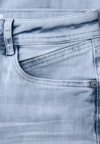 Zbliżenie na jasnoniebieskie dżinsy z denimu, pokazujące przednią kieszeń, szlufkę na pasek, detale szwów oraz metalowy nit po prawej stronie.