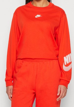 Personne portant un haut manches longues Nike orange vif avec un logo blanc sur la poitrine et la manche, assorti à un pantalon orange.