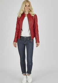 Rote Lederjacke mit Stehkragen, Reißverschlüssen und rautenförmigen Steppakzenten, kombiniert mit dunklen Jeans und weißen Sneakern.