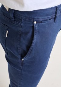 Tmavě modré džínové kalhoty se hladkým povrchem, obsahující přední kapsu a kovové akcenty. Pas má poutka na pásek.