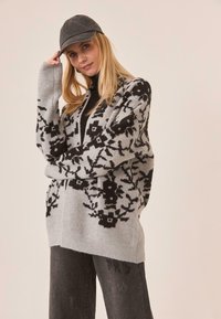 Cárdigan gris con un patrón que presenta dise�ños florales en negro, de manga larga y corte relajado. Combinado con un cuello alto negro y jeans grises.