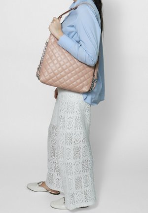 Woman wearing a light blue blouse, white lace pants, white slip-on shoes, holding a large beige quilted handbag with chain straps.
