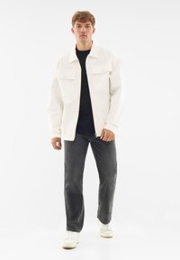 White denim jacket with buttoned chest pockets, black t-shirt underneath, dark jeans, and white sneakers against a plain background.