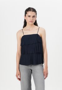 Top plissé bleu marine avec des volants superposés, bretelles spaghetti ajustables et une silhouette fluide. Assorti à un jean large gris clair.