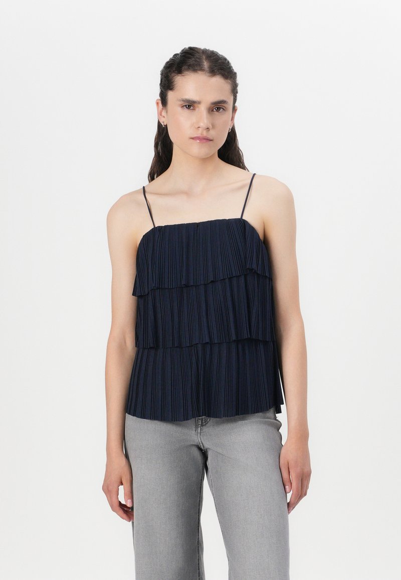 Top plissé bleu marine avec des volants superposés, bretelles spaghetti ajustables et une silhouette fluide. Assorti à un jean large gris clair.