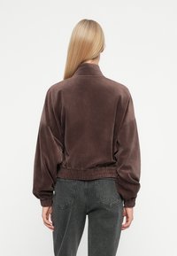 Smešna smeđa bomber jakna s visokim ovratnikom, elastičnom strukom i širokim rukavima, u kombinaciji s tamno sivim farmerkama.