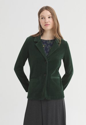 Woman wearing a dark green corduroy blazer over a floral top and dark skirt, standing with hands behind her back on a plain light background.