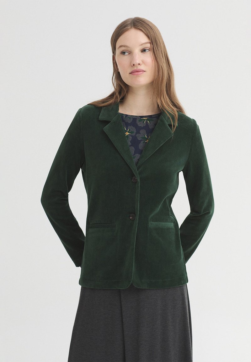 Femme portant un blazer en velours côtelé vert foncé sur un top fleuri et une jupe sombre, debout les mains dans le dos sur un fond clair uni.