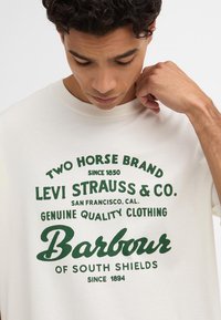 Kreemivärvi puuvillane t-särk, millel on roheline tekstigraafika, mis sisaldab brändi nime ja asukoha üksikasju. Ümar kaelus ja lühikesed varrukad.