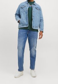 Vaaleansininen farkkutakki kauluksella, kahdella rintataskulla, päällä vihreä t-paita. Siniset farkut ja valkoiset lenkkarit täydentävät asun.