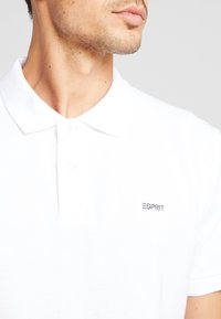 Vit pikétröja i bomull, med klassisk krage, treknappsstängning och en liten marinblå logo på vänster bröst.