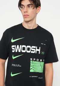 Črna majica s kratkimi rokavi, ki vsebuje drzne bele napise "SWOOSH", zelene Nike logotipe in grafične podrobnosti v strukturirani zasnovi. Narejena iz bombaža, s sproščenim krojem.