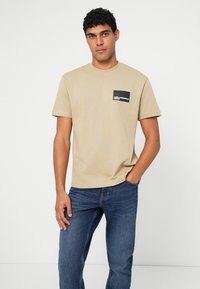 Bežinis trumparankovis marškinėliai su apvalia iškirpte, turintis juodą kišenę su balta logotipo užrašu, derinamas su mėlynomis džinsomis. Lygus medvilnės audinys.