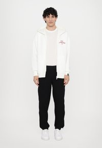 Biela zip-up mikina s červeným textom, vrstvená nad svetložltým tričkom, v kombinácii s čiernymi teplákmi a bielymi teniskami. Hladká textura, voľný strih.
