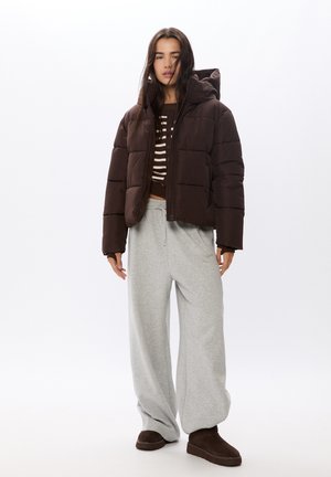 Braune Pufferjacke mit Kapuze, graue oversized Hosen und dunkelbraune Hausschuhe. Gestreiftes Oberteil unter der Jacke. Glatte Textur.