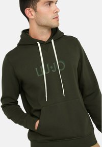 Donkergroene capuchontrui met voorvak, witte trekkoorden en een subtiel gedrukt logo op de borst. Gemaakt van een zachte katoenmengsel.