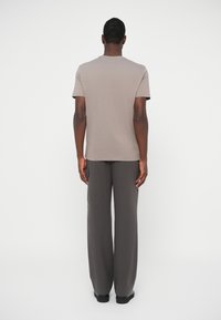 Man standing with back to camera wearing beige short-sleeve T-shirt and dark gray wide-leg pants with side pocket against white background.