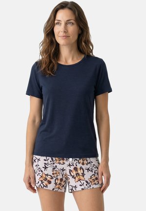 Kobieta z falującymi brązowymi włosami, ubrana w granatowy t-shirt z krótkim rękawem i jasne, kwiatowe szorty, stojąca na jednolitym tle.