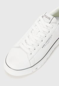 White leather sneaker with textured surface, round toe, and flat laces. Features branding near the tongue and a thin black accent around the sole.