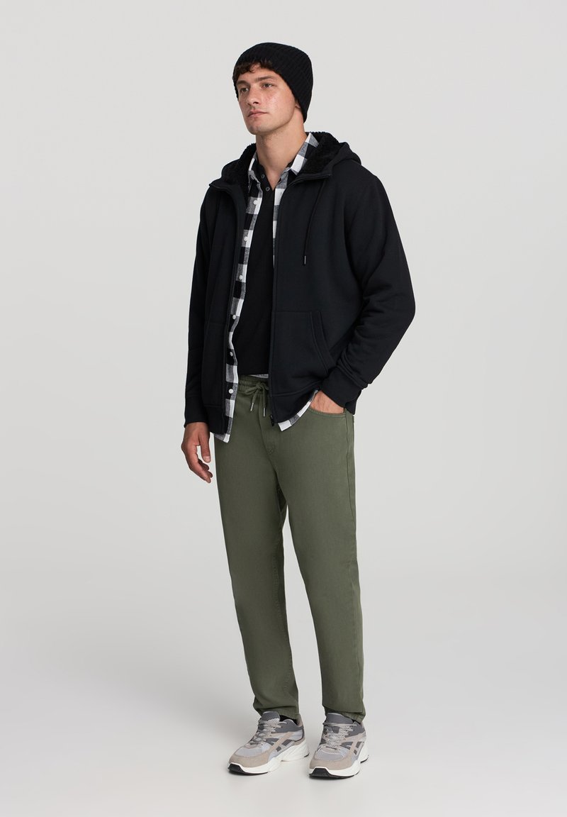 Uomo che indossa un berretto nero, una felpa nera con zip, una camicia a scacchi bianca e nera, una maglietta nera, pantaloni verde oliva e sneakers grigie in piedi.