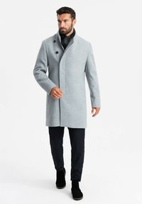 Abrigo de lana en mezcla gris claro con cuello alto, botones dobles y una silueta estilizada, combinado con pantalones oscuros y botas negras.