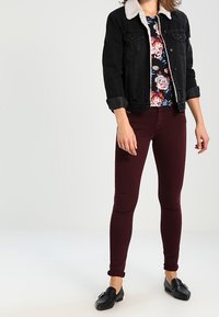 Schwarze Jeansjacke mit weißem Fleece-Kragen, blumiges Oberteil, bordeauxrote enge Jeans und schwarze Ballerinas, wodurch ein lässiges Outfit entsteht.