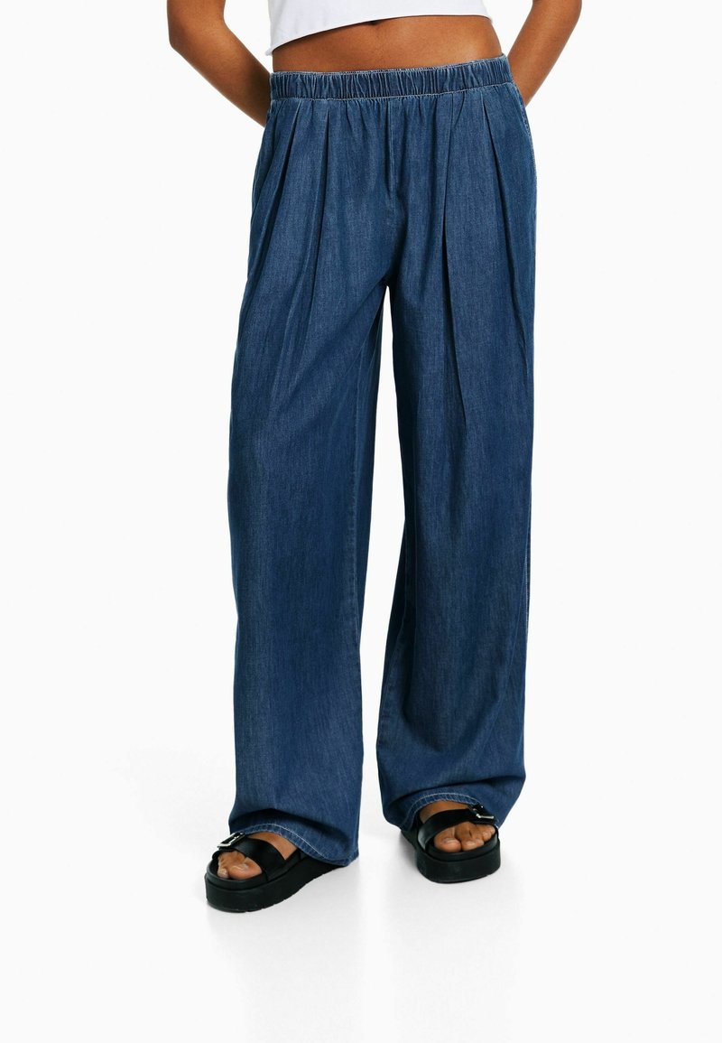 Pantalones vaqueros de pierna ancha en azul oscuro con cintura elástica, que presentan pliegues frontales y una textura suave. Combinados con sandalias negras.